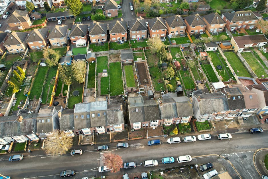 An aerial view of a residential area showing multiple back gardens separated by wooden fences along a suburban street. The gardens feature well-maintained lawns with neatly trimmed grass, flower beds